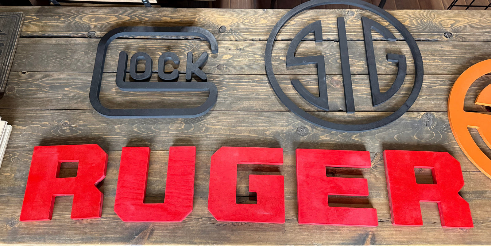 A rustic wooden sign featuring raised metal lettering. The left side has large red letters spelling 'RUGER' vertically, while the right side has black metal logo-style shapes, including a central 'RUGER' emblem and circular and square monogram designs.